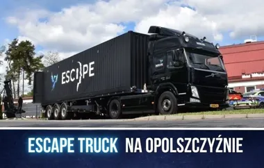 Czarny ciężarówka z napisem ESCAPE na naczepie stoi na drodze w słoneczny dzień, w tle budynek z czerwonym dachem.