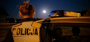 Policjantka w kamizelce z napisem POLICJA stoi przy radiowozu z włączonym błękitnym światłem nocą pod pełnią księżyca.