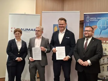 Cztery osoby w garniturach stoją podczas uroczystości podpisania dokumentu, w tle roll-upy firmy Baumap i Szpitala Wojewódzkiego w Opolu.